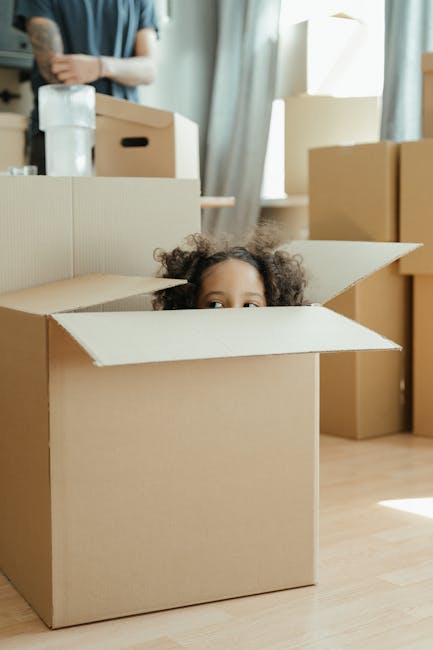 A young child with curly hair is inside an open cardboard moving box, with only the child's head and a small part of their face visible peeking above the flaps. The box is placed on a light wooden floor within a bright room filled with natural light, with several other closed cardboard boxes of various sizes arranged around. In the background, a person stands near a window with sheer curtains, holding a rolled-up item wrapped in plastic, and surrounded by more packed boxes, some labeled with black marker. Moving equipment such as a small hand trolley, packing materials including bubble wrap and plastic wrapping, and furniture like a chair is visible nearby. This scene depicts the packing and home relocation process, with the setting emphasizing the careful handling of household items during furniture transport and loading for a house removal service provided by Man with Van Selhurst, supporting the topic of transparent Selhurst removals pricing.