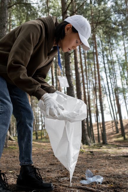 A woman dressed in casual outdoor clothing, including a white cap, brown hoodie, and white gloves, is seen bending down in a forested area with tall, slender trees and a dirt ground. She is holding a white plastic garbage bag open with one hand while using the other hand to pick up a discarded plastic water bottle from the ground, which appears to be part of a litter collection effort. The scene captures a cleanup activity, emphasizing environmentally responsible behaviour during home relocation or moving logistics. The woman is engaged in removing waste from the outdoor environment, supporting efforts related to packing or disposing of unwanted items before transportation. Natural light filters through the trees, illuminating the scene, which is associated with responsibilities often undertaken by professional removals services such as Man with Van Selhurst.