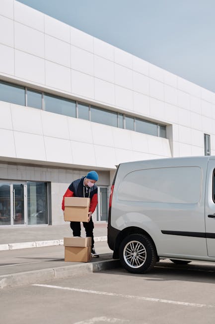 A man with a beard, wearing a white t-shirt, blue jeans, and a black cap, is seen loading three medium-sized cardboard boxes onto the side of a white panel van parked on the street in front of residen