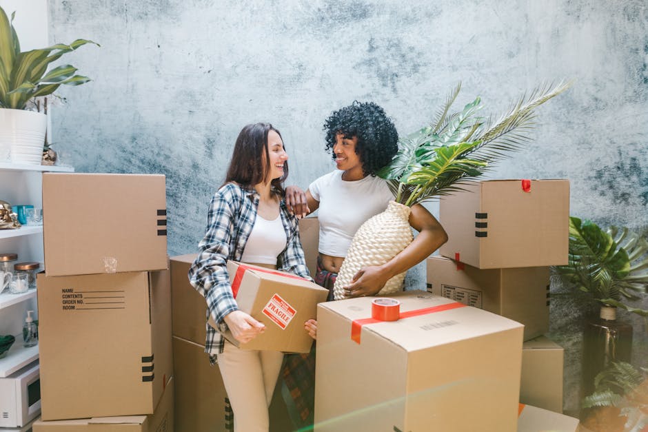 Two women engaged in a home relocation process indoors, surrounded by stacks of cardboard boxes sealed with red tape, some labeled with details such as 'name,' 'contents,' and 'room no.' One woman has straight, shoulder-length dark hair and is dressed in a plaid shirt and light-colored pants, holding a small box with a red 'fragile' sticker. The other woman has curly black hair and is wearing a white t-shirt, holding a woven basket with large green palm leaves inside. They are smiling and standing close together, with a concrete-textured wall in the background and some furniture and decorative items like plants on shelves visible on the left side. The image captures the loading process of packing and moving essentials as part of a professional removals service, with a focus on careful handling and packing during a house move facilitated by Man with Van Selhurst.