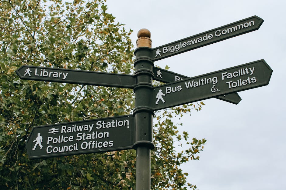 A black multi-directional street signpost standing outdoors against a backdrop of green leafy trees and a clear, slightly overcast sky. The signpost features several rectangular signs with white text and icons indicating directions to local amenities, including a library, railway station, police station, council offices, Biggleswade Common, bus waiting facility, and toilets with accessible options. The signs are mounted on a central pole with protective brackets, and each sign points in a different direction. This scene illustrates typical signage used in residential areas or town centres to guide pedestrians and visitors, relevant to house removals and local relocation services offered by Man with Van Selhurst, especially in relation to planning and navigating moving logistics in the Croydon area.