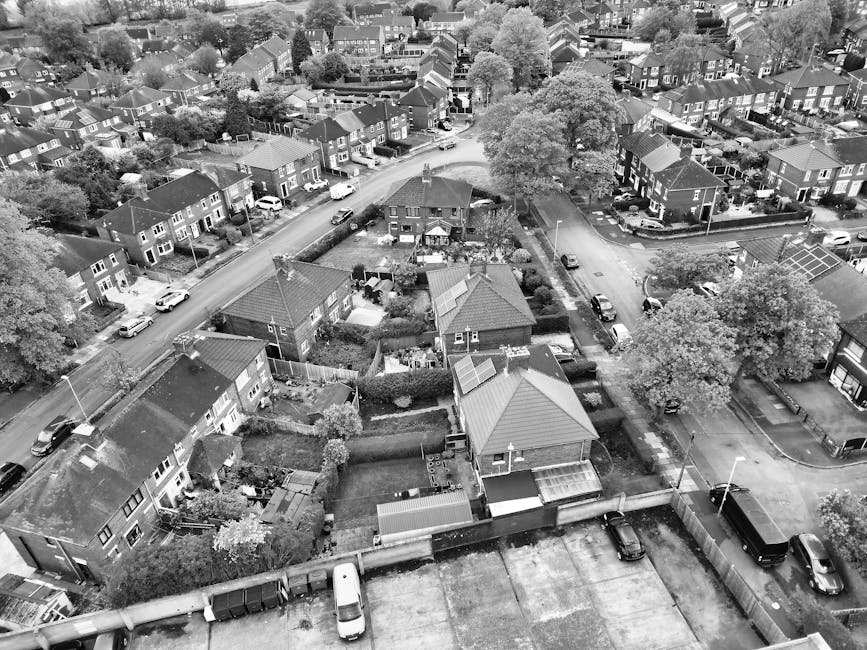 An aerial view of a residential neighbourhood showing multiple detached and semi-detached houses with pitched roofs, surrounded by trees and greenery. Several parked cars are visible along the streets and in driveways. In the foreground, a house with a fenced garden contains various objects, including outdoor furniture and garden sheds. A van is parked on the street near the house, and a large tree provides shade over part of the area. The scene captures a typical suburban setting during daylight, with signs of ongoing home relocation or furniture transport, as part of the moving process involving Man with Van Selhurst, who specialise in removals within the Croydon area.