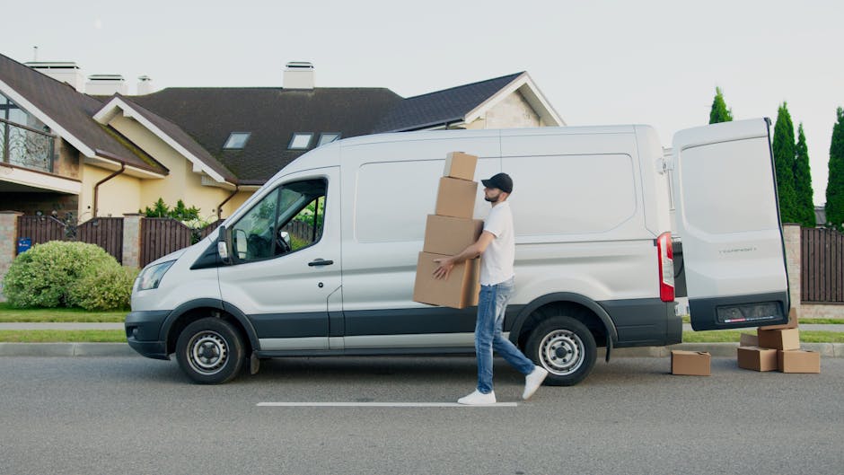 A young man with a serious expression, wearing a black cap and a white polo shirt, stands inside the open rear of a moving van during a home relocation. Behind him, the van's interior is filled with s