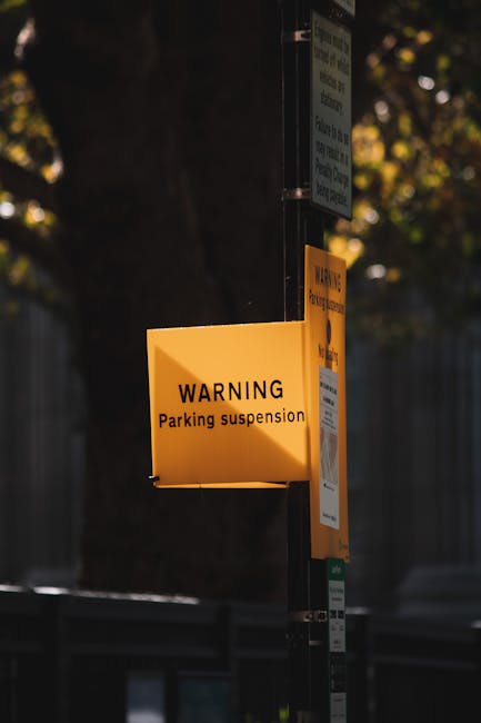 A yellow warning sign attached to a black metal pole indicating 'Parking suspension' beneath a larger informational sign, with a background of blurred trees and a fence. The sign is positioned on a pavement near a property, suggesting restrictions relevant to house removals and moving logistics. The area is well-lit, highlighting the text and the sign's visibility for passing vehicles and pedestrians involved in furniture transport or home relocation activities. The scene reflects the typical signage encountered during organised removals, where parking restrictions facilitate the loading and unloading process for vehicles such as vans or trucks used by Man with Van Selhurst when assisting with house moves, packing, and furniture transport.