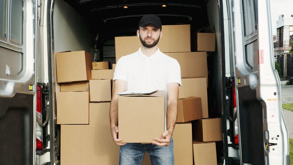 Inside a white moving van, a man wearing a black cap, black face mask, yellow vest, and gloves is seated in the driver’s seat, holding a clipboard or document in his right hand. The van’s dashboar
