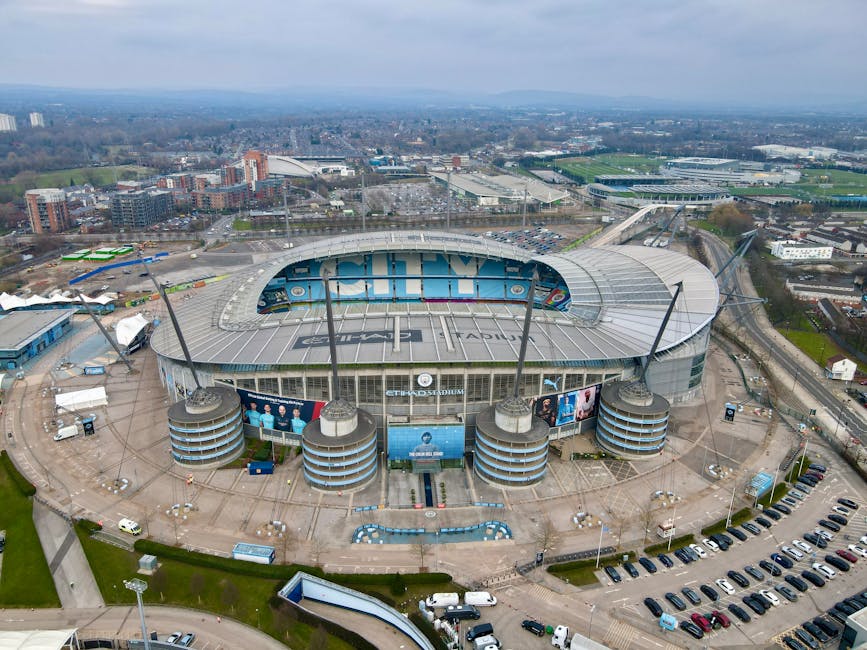 An aerial view of a large football stadium with a partially transparent roof covering the pitch and seating areas, surrounded by parking lots filled with cars and adjacent roads. The stadium's exterio