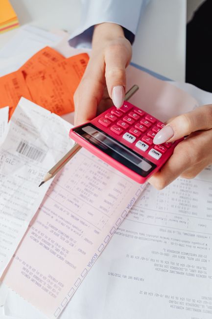 Close-up of a person's hands holding a pink calculator and working with various paperwork, receipts, and documents on a table. The papers include printed invoices, packing lists, and printed forms, so