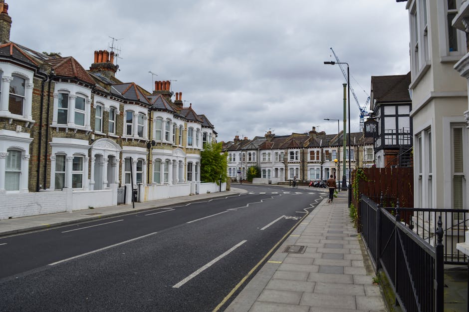 A residential street scene captured in daylight under overcast skies, featuring a row of Victorian-style terraced houses with white facades, bay windows, and red-tiled roofs. The street is lined with a paved sidewalk on the right, separated from the road by a metal railing, with a person walking along the pavement. In the foreground, a black van associated with Man with Van Selhurst is parked, likely involved in a home relocation or furniture transport process. The scene includes various cardboard boxes and packing materials placed near the entrance of one of the houses, indicating packing and moving activities typical of professional removals services. Several street lights and cranes are visible in the background, with some buildings under construction or renovation, reflecting a typical urban environment suitable for local moves along Selhurst Road to South Norwood; all elements point to professional loading, packing, and transportation operations coordinated by [COMPANY_NAME].