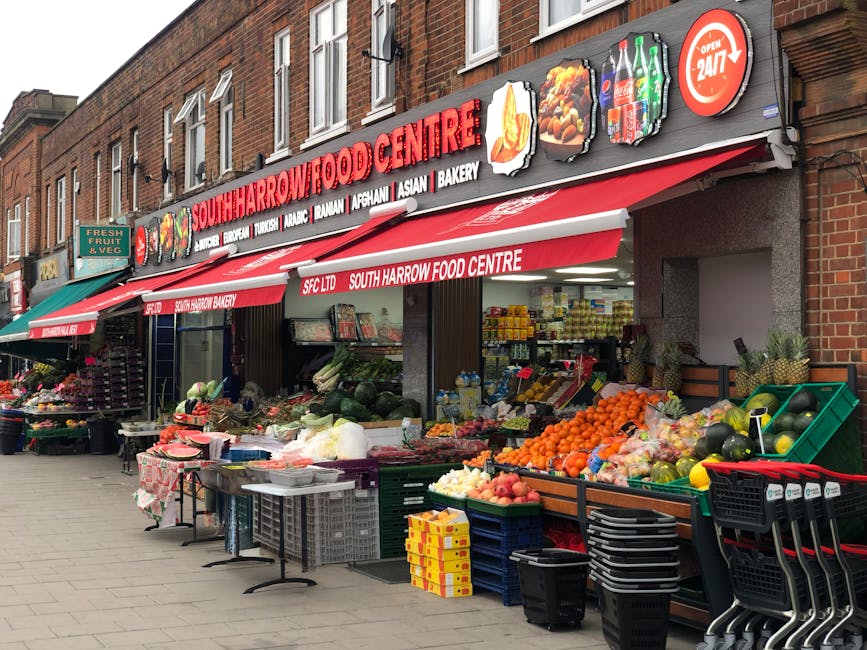 A residential street scene captured in daylight under overcast skies, featuring a row of Victorian-style terraced houses with white facades, bay windows, and red-tiled roofs. The street is lined with a paved sidewalk on the right, separated from the road by a metal railing, with a person walking along the pavement. In the foreground, a black van associated with Man with Van Selhurst is parked, likely involved in a home relocation or furniture transport process. The scene includes various cardboard boxes and packing materials placed near the entrance of one of the houses, indicating packing and moving activities typical of professional removals services. Several street lights and cranes are visible in the background, with some buildings under construction or renovation, reflecting a typical urban environment suitable for local moves along Selhurst Road to South Norwood; all elements point to professional loading, packing, and transportation operations coordinated by [COMPANY_NAME].