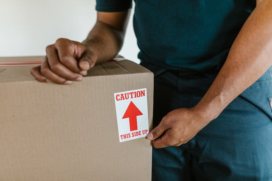 A person wearing a dark green t-shirt and dark pants is in the process of carefully handling a large cardboard moving box during a home relocation. The individual is gripping the box firmly with one h
