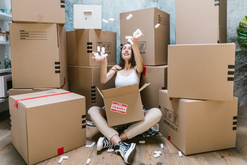 A person wearing a dark green t-shirt and dark pants is in the process of carefully handling a large cardboard moving box during a home relocation. The individual is gripping the box firmly with one h